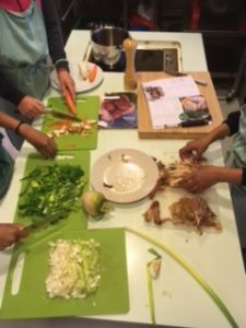 Bord med kokeplate, kasserolle, tallerken og fire skjærefjeler med oppkuttede grønnsaker. Hender som holder kniver sees i billedkanten.